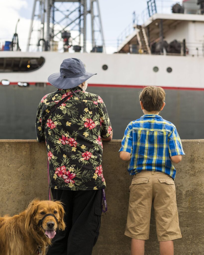 Two people watching a boat with a dog looking at camera.
